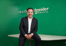 Schneider Electric: Del respaldo energético al monitoreo inteligente; cómo ampliar el valor del canal en infraestructura crítica Fotografía de plano medio de José Alberto Llavot posando sentado de lado sobre el borde de una mesa blanca, vistiendo traje ejecutivo oscuro y camisa blanca sin corbata, sonriendo frente a una pared verde institucional con el logotipo "Life Is On Schneider Electric" en letras blancas.