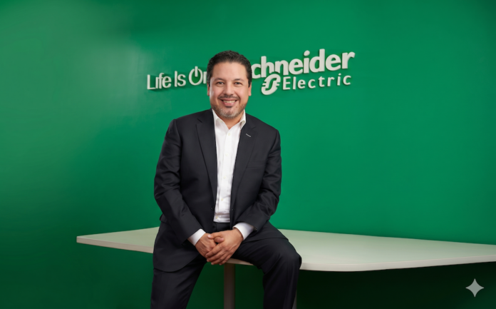 Fotografía de plano medio de José Alberto Llavot posando sentado de lado sobre el borde de una mesa blanca, vistiendo traje ejecutivo oscuro y camisa blanca sin corbata, sonriendo frente a una pared verde institucional con el logotipo 