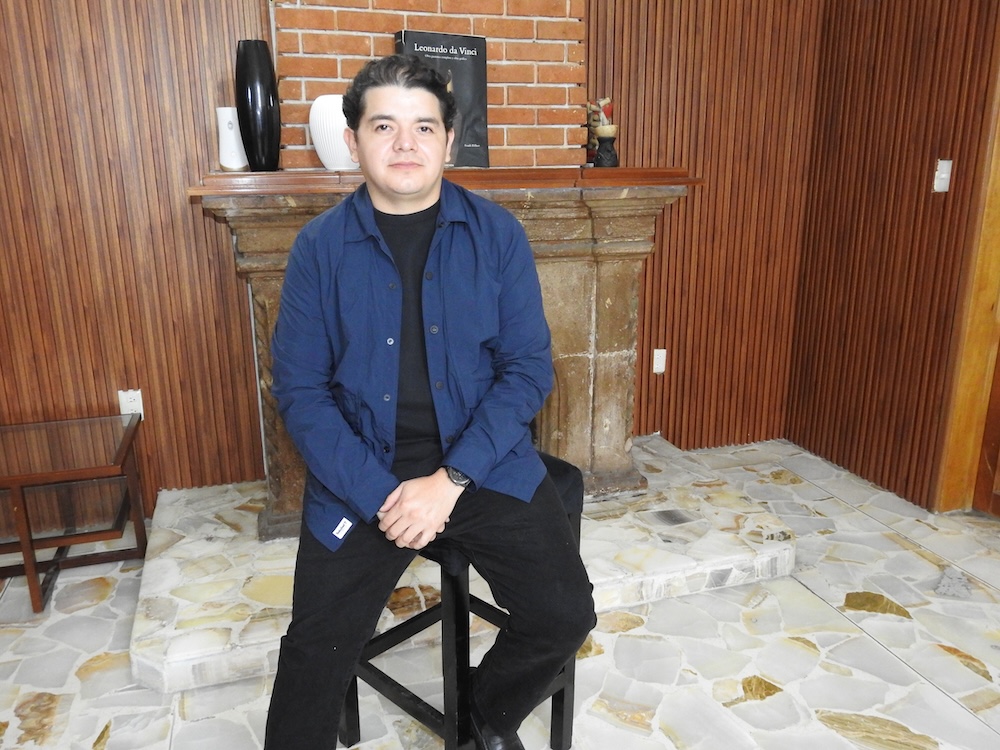 Fotografía de retrato de José Luis Mejía sentado sobre un taburete alto en una sala con paredes con paneles de madera, vistiendo una sobrecamisa azul marino sobre una playera negra.