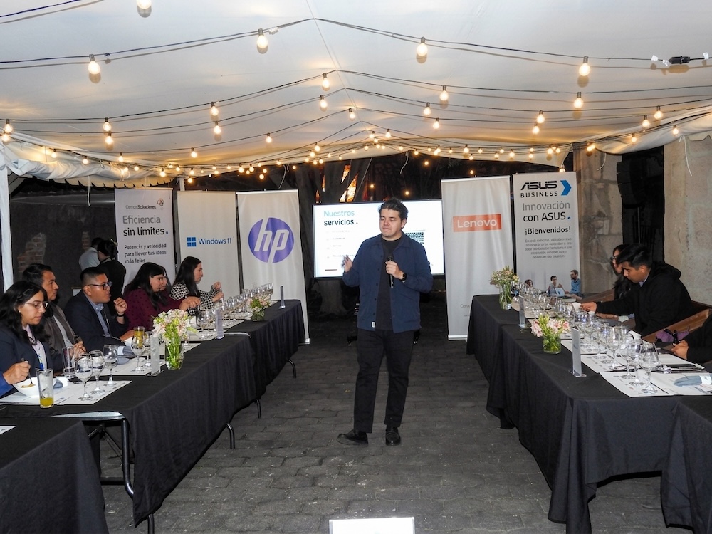 Fotografía de José Luis Mejía hablando con un micrófono en el centro de una terraza acondicionada con mesas para cena. A sus espaldas se observan mamparas publicitarias de marcas como HP, Lenovo y ASUS Business.