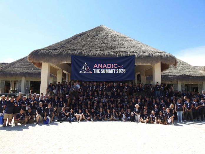 Fotografía de un grupo multitudinario de personas posando alegremente sobre la arena blanca de una playa, frente a una gran estructura arquitectónica tipo palapa. La inmensa mayoría de los asistentes viste camisas tipo polo en color azul oscuro y, en el centro de la formación, sostienen una gran lona azul con el logotipo y el texto 