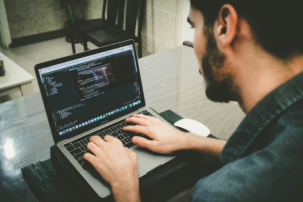 Fotografía en ángulo posterior de un hombre joven sentado frente a un escritorio de madera, escribiendo en el teclado de una computadora portátil donde se observa un entorno de desarrollo integrado con líneas de código de programación.