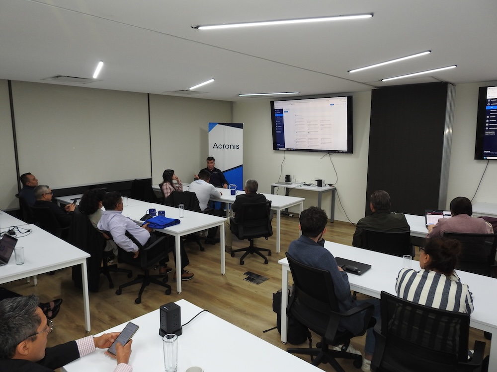 Fotografía en ángulo picado de una sala de capacitación corporativa donde un grupo de profesionales e integradores de TI escuchan a un ponente ubicado al fondo, quien realiza una presentación en pantalla sobre Acronis Cyber Protect Cloud.