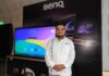 BenQ transforma la educación en México con innovación tecnológica e interactiva Fotografía en plano medio de Mauricio Molina, Sell Out Manager en BenQ Latam, vistiendo una guayabera blanca con bordados tradicionales y anteojos, sonriendo al posar frente a una pantalla interactiva encendida y un panel publicitario negro de la marca en un evento tecnológico.