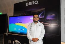 BenQ transforma la educación en México con innovación tecnológica e interactiva Fotografía en plano medio de Mauricio Molina, Sell Out Manager en BenQ Latam, vistiendo una guayabera blanca con bordados tradicionales y anteojos, sonriendo al posar frente a una pantalla interactiva encendida y un panel publicitario negro de la marca en un evento tecnológico.