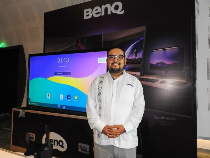 Fotografía en plano medio de Mauricio Molina, Sell Out Manager en BenQ Latam, vistiendo una guayabera blanca con bordados tradicionales y anteojos, sonriendo al posar frente a una pantalla interactiva encendida y un panel publicitario negro de la marca en un evento tecnológico.