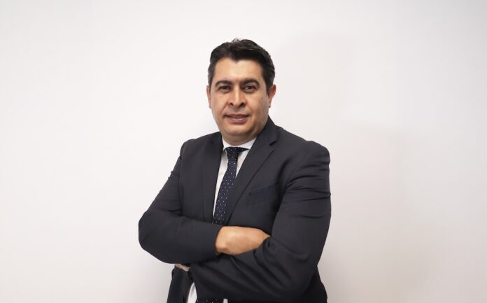 Miguel Espinoza asume la dirección interina de Grupo CVA Fotografía de retrato corporativo de Miguel Espinoza posando de pie con los brazos cruzados y mirando al frente con una ligera sonrisa. Viste un traje formal de color azul marino, camisa blanca y corbata oscura con discreto patrón de lunares blancos, frente a un fondo blanco liso de estudio.