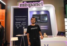 Mixpanel impulsa el análisis de productos digitales en Latam Fotografía de un representante de Mixpanel vistiendo una camiseta negra con el logotipo de la marca, posando de pie y sonriendo frente a un stand corporativo iluminado con luces moradas de neón. En el fondo, un panel muestra el lema "Convirtiendo datos en decisiones" junto a logotipos de diversas empresas, mientras una pantalla exhibe un tablero de análisis de rendimiento.