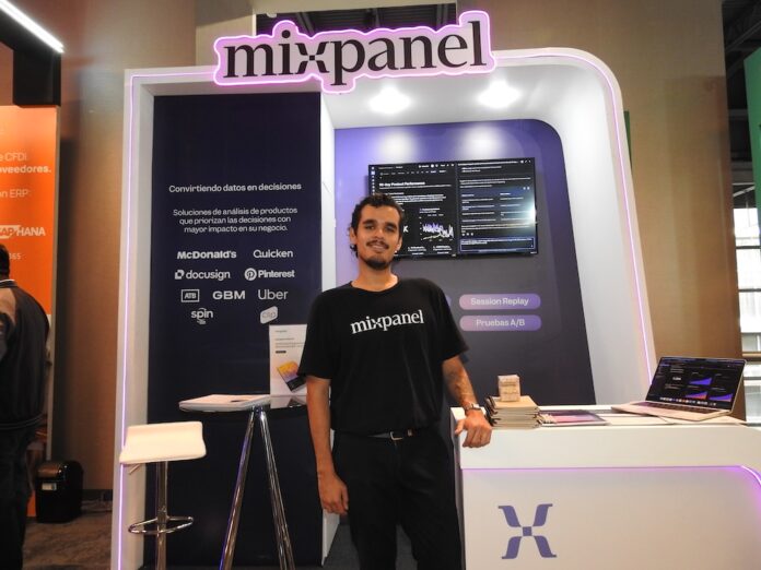 Mixpanel impulsa la adopción de analítica de productos digitales en México Fotografía de un representante de Mixpanel vistiendo una camiseta negra con el logotipo de la marca, posando de pie y sonriendo frente a un stand corporativo iluminado con luces moradas de neón. En el fondo, un panel muestra el lema
