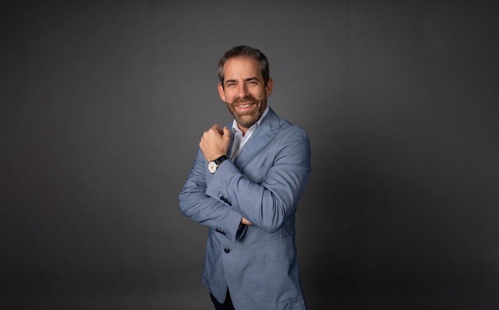 Fotografía de retrato de Moisés Avelar posando de medio cuerpo con los brazos cruzados y sonriendo hacia la cámara. Viste un saco azul claro sobre una camisa blanca sin corbata, destacando un reloj con correa de cuero en su muñeca izquierda, frente a un fondo neutro de color gris oscuro.