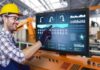 ViewSonic lanza en América Latina su nueva línea exclusiva de monitores táctiles industriales Open Frame Fotografía de un operador industrial con casco amarillo, lentes de protección y guantes negros de trabajo, sonriendo mientras toca con el dedo un panel HMI de automatización industrial que despliega esquemas de líneas de producción y gráficos de rendimiento.