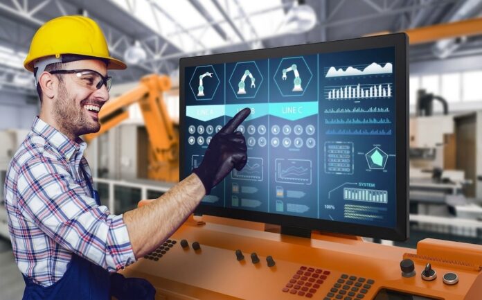 Monitores táctiles industriales ViewSonic Open Frame para automatización y plantas de producción Fotografía de un operador industrial con casco amarillo, lentes de protección y guantes negros de trabajo, sonriendo mientras toca con el dedo un panel HMI de automatización industrial que despliega esquemas de líneas de producción y gráficos de rendimiento.