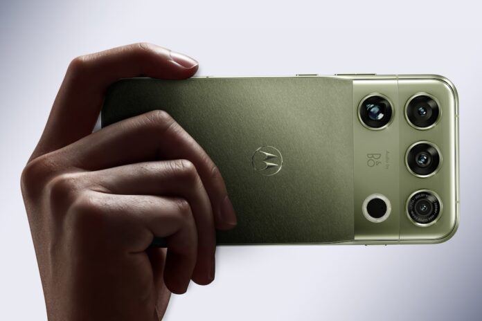 Fotografía en plano detalle sosteniendo horizontalmente un smartphone motorola signature 27 en acabado PANTONE Capulet Olive con textura textil cepillada, mostrando la insignia icónica de Motorola y el módulo de cámaras con el grabado 