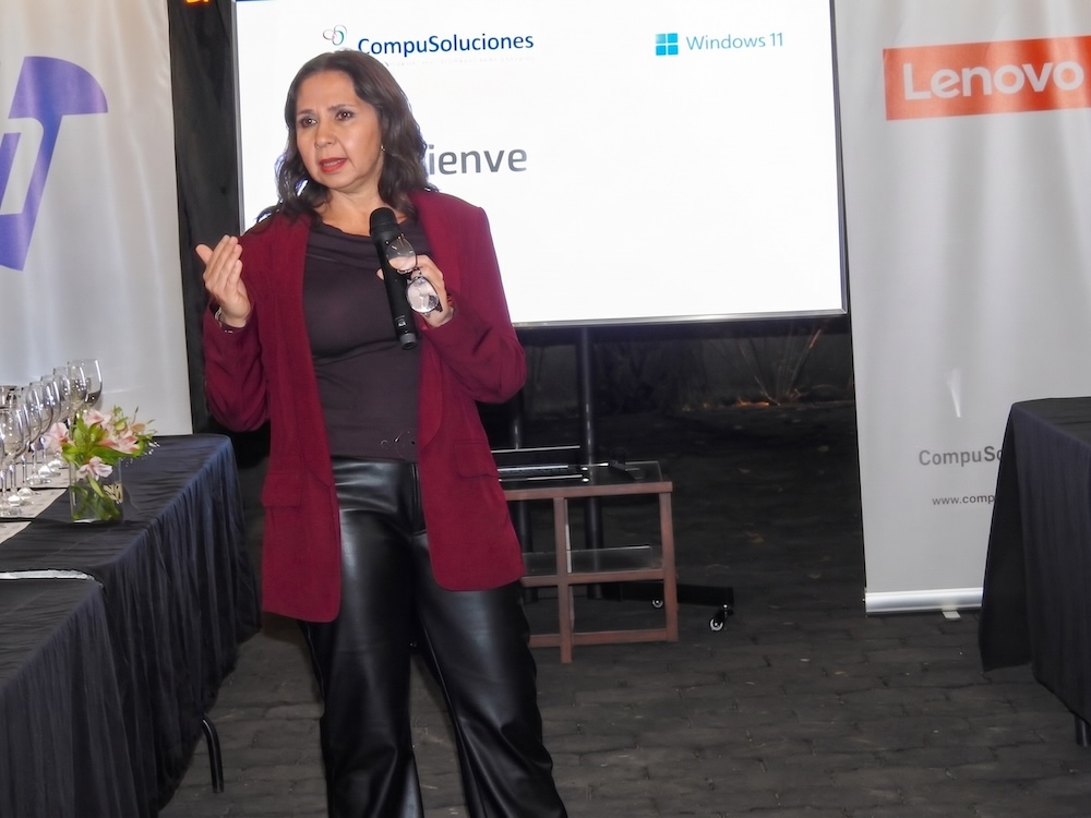 Fotografía de Adriana Valdez de pie sosteniendo un micrófono mientras imparte una conferencia. Viste un saco color guinda sobre una blusa oscura y pantalones de piel, con un proyector y mamparas de marcas al fondo.