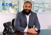 Eaton y la revolución del espacio de trabajo: energía y resiliencia digital Retrato de plano medio del ejecutivo Ramón León, sonriendo a la cámara y sentado detrás de un escritorio de oficina con un teléfono IP de color oscuro en primer plano. Viste un saco de vestir a cuadros grises y azules sobre una camisa blanca de cuello abierto. Al fondo se observa un ventanal con una vista panorámica de la Ciudad de México y, en la esquina superior izquierda, el logotipo de Eaton con la leyenda "Powering Business Worldwide".