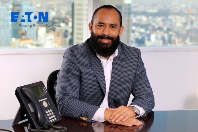 Retrato de plano medio del ejecutivo Ramón León, sonriendo a la cámara y sentado detrás de un escritorio de oficina con un teléfono IP de color oscuro en primer plano. Viste un saco de vestir a cuadros grises y azules sobre una camisa blanca de cuello abierto. Al fondo se observa un ventanal con una vista panorámica de la Ciudad de México y, en la esquina superior izquierda, el logotipo de Eaton con la leyenda 