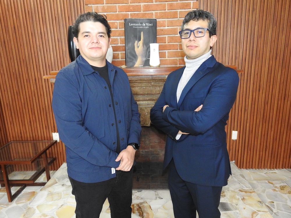 Fotografía de tres cuartos de cuerpo que muestra a Carlos Enrique García y José Luis Mejía posando sonrientes en una sala con paredes de madera, frente a una chimenea decorativa de ladrillo.