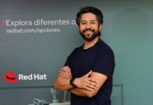 Red Hat: El arquitecto del futuro del trabajo y la nube híbrida sin ataduras Fotografía de retrato de Roberto Calva posando sonriente con los brazos cruzados en un tono casual con camiseta oscura, frente a una pared de fondo verde grisáceo decorada con el logotipo corporativo de Red Hat y la leyenda publicitaria "redhat.com/opciones".