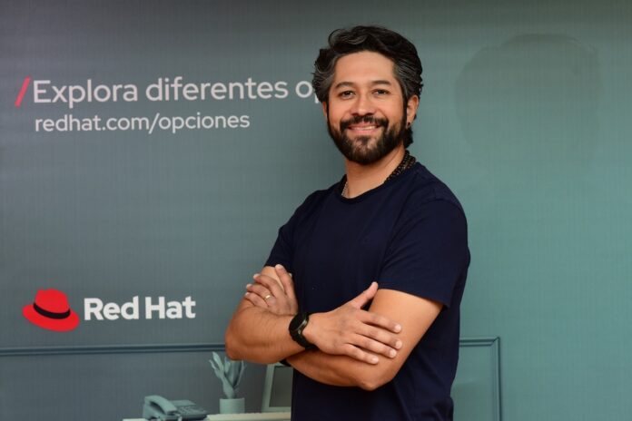 Fotografía de retrato de Roberto Calva posando sonriente con los brazos cruzados en un tono casual con camiseta oscura, frente a una pared de fondo verde grisáceo decorada con el logotipo corporativo de Red Hat y la leyenda publicitaria 