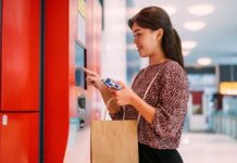 SONDA potencia el retail en México con autocobro inteligente y tecnología híbrida Fotografía de plano medio que muestra a una mujer joven sonriendo mientras interactúa con la pantalla táctil de un quiosco de autocobro de color rojo en una tienda. La mujer sostiene su teléfono inteligente con una mano y lleva una bolsa de papel kraft colgada del brazo en un entorno comercial moderno y bien iluminado.
