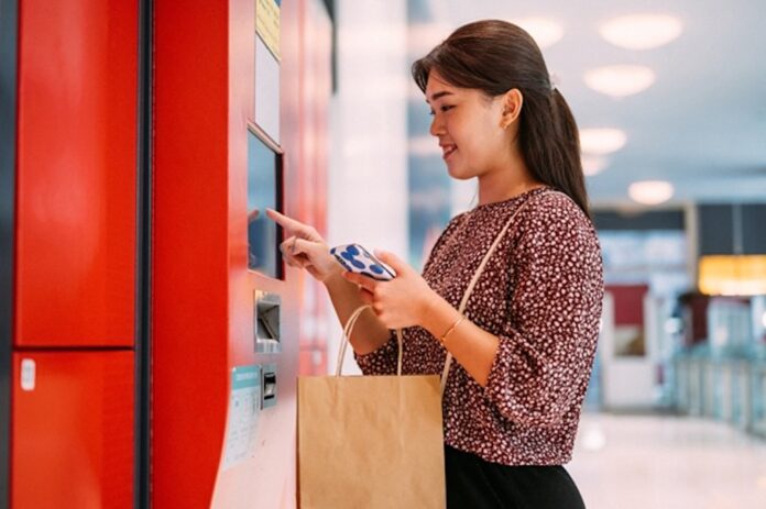 Fotografía de plano medio que muestra a una mujer joven sonriendo mientras interactúa con la pantalla táctil de un quiosco de autocobro de color rojo en una tienda. La mujer sostiene su teléfono inteligente con una mano y lleva una bolsa de papel kraft colgada del brazo en un entorno comercial moderno y bien iluminado.
