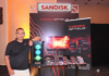 El poder de Sandisk revoluciona el almacenamiento para gamers y creadores Fotografía en plano medio de Alejandro Medina, especialista en marketing y ventas de SanDisk, recargado sonriente junto a una torre de computadora con iluminación RGB en el estand de lanzamiento de la línea SanDisk Optimus.
