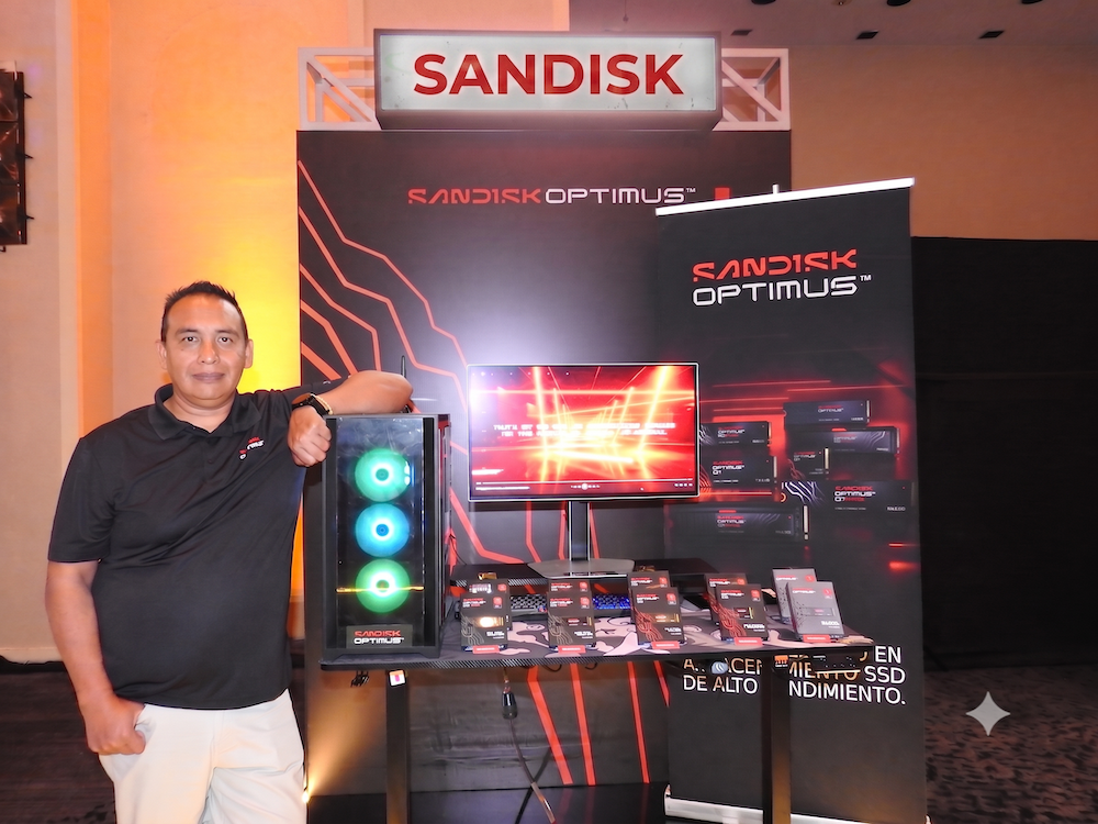 SanDisk presenta la línea de unidades de estado sólido Optimus en el AMD Summit 2026 Fotografía en plano medio de Alejandro Medina, especialista en marketing y ventas de SanDisk, recargado sonriente junto a una torre de computadora con iluminación RGB en el estand de lanzamiento de la línea SanDisk Optimus.