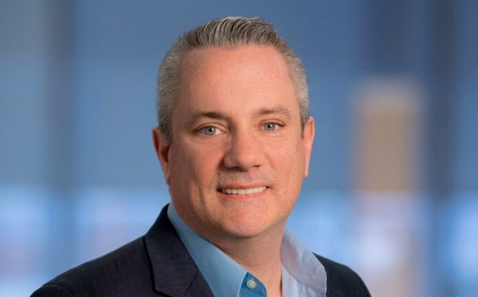 Fotografía de retrato corporativo de Scott Barlow, Vicepresidente de Sophos, luciendo un saco oscuro y camisa azul claro sobre un fondo de estudio desenfocado.