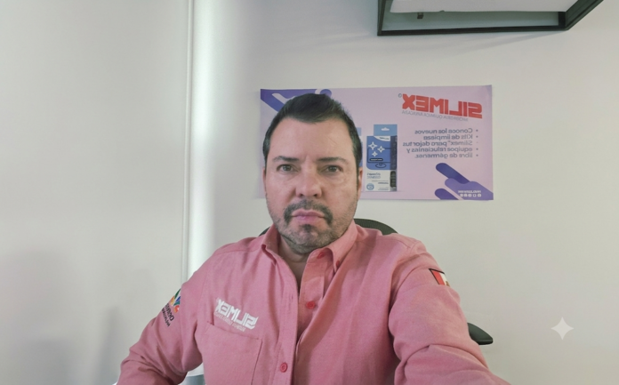 Fotografía de retrato de Alfredo Álvarez Buylla, Gerente de ventas de Silimex. Aparece mirando directamente a la cámara, vistiendo una camisa de tono rosa salmón con el logotipo de la marca bordado en el pecho derecho. Al fondo, sobre una pared blanca, se observa un póster publicitario de los productos y kits de limpieza de la compañía.
