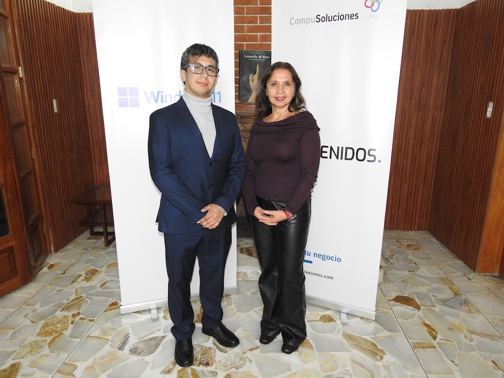 Sinergia entre CompuSoluciones y Microsoft para transformar el canal en México Fotografía de plano entero que muestra a Carlos Enrique García y Adriana Valdez posando juntos de pie sobre un piso de piedra clara, frente a mamparas publicitarias de Windows 11 y CompuSoluciones.