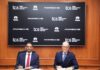 TCS y Porsche se unen para transformar la movilidad inteligente mediante IA y la adquisición de MHP Fotografía institucional de K. Krithivasan y el Dr. Michael Leiters sentados en una mesa de reuniones de madera con bolígrafos en la mano para la firma de un documento corporativo. En el fondo se aprecia una pantalla que muestra repetidamente los logotipos oficiales de Tata Consultancy Services, TATA y Porsche.