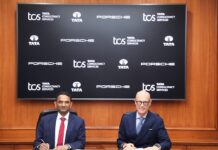 TCS y Porsche se unen para transformar la movilidad inteligente mediante IA y la adquisición de MHP Fotografía institucional de K. Krithivasan y el Dr. Michael Leiters sentados en una mesa de reuniones de madera con bolígrafos en la mano para la firma de un documento corporativo. En el fondo se aprecia una pantalla que muestra repetidamente los logotipos oficiales de Tata Consultancy Services, TATA y Porsche.
