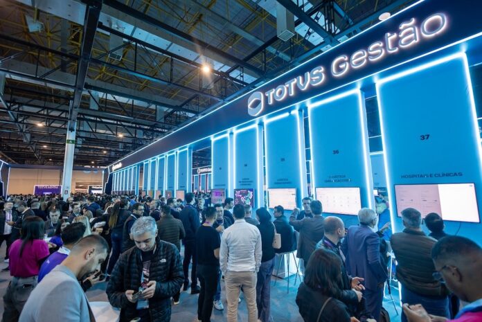 Profesionales y ejecutivos de tecnología recorren los stands de demostración e interacción durante el encuentro tecnológico en Expo Center Norte. Fotografía en ángulo panorámico de la concurrida área de exhibición tecnológica en Universo TOTVS, iluminada con letreros neón azules donde destaca la marca