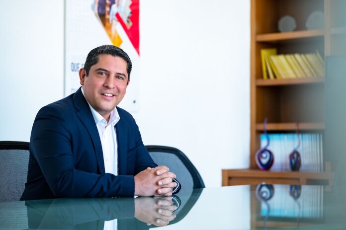 Fotografía de retrato corporativo en plano medio de Víctor Manuel Urzúa, subdirector comercial de CompuSoluciones, sonriendo con las manos entrelazadas sobre un escritorio de cristal en su oficina.