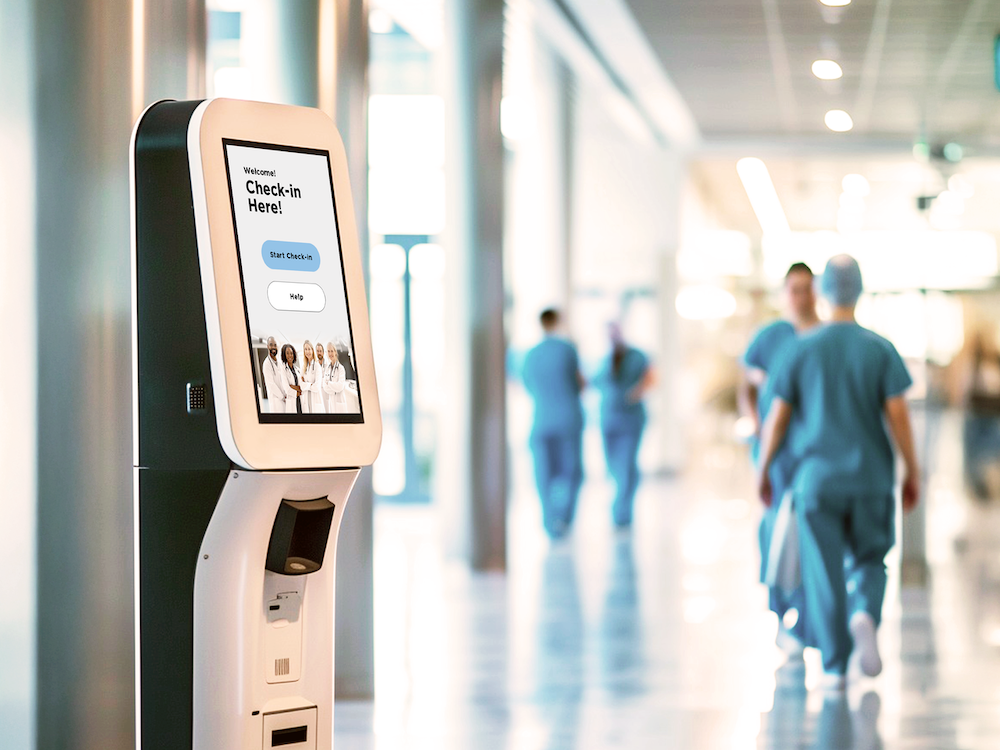 Fotografía de un quiosco de autoservicio de color blanco y negro instalado en el pasillo de un centro médico u hospital, con una pantalla táctil vertical que despliega una interfaz de registro de pacientes ("Check-in Here!"), mientras personal de salud con vestimenta médica camina desenfocado en segundo plano.