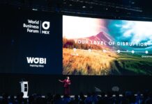 WOBI analiza la paradoja de la IA en México donde el 55% de las empresas la usa pero solo el 6% crea valor real Fotografía en perspectiva abierta del escenario principal de un auditorio corporativo donde una ponente vestida de traje rojo gesticula frente a una audiencia multitudinaria. En el fondo se aprecia una pantalla gigante con el logotipo del World Business Forum México de WOBI y la leyenda "YOUR LEVEL OF DISRUPTION" acompañada de un gráfico de escala conceptual entre un estado calmo y uno caótico.