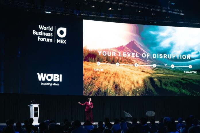 WOBI analiza el impacto estratégico de la inteligencia artificial y el futuro del trabajo en el World Business Forum México 2026 Fotografía en perspectiva abierta del escenario principal de un auditorio corporativo donde una ponente vestida de traje rojo gesticula frente a una audiencia multitudinaria. En el fondo se aprecia una pantalla gigante con el logotipo del World Business Forum México de WOBI y la leyenda