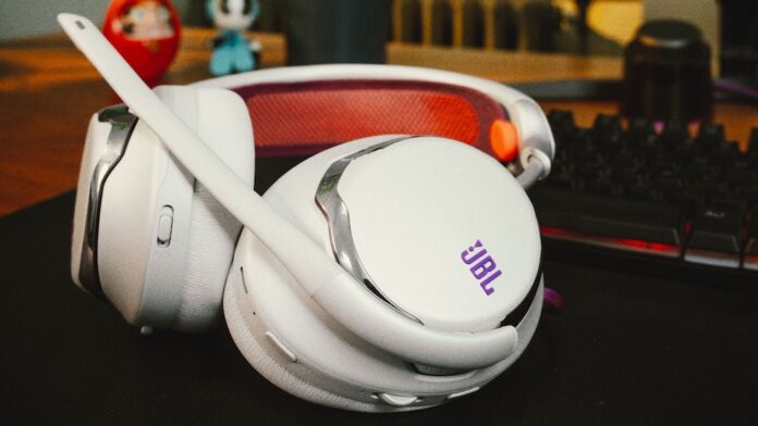 Fotografía en plano detalle de los audífonos inalámbricos para videojuegos JBL Quantum 950 en color blanco con acentos morados y naranjas, mostrando su micrófono boom, la diadema de malla transpirable y las almohadillas descansando sobre un escritorio junto a un teclado mecánico.
