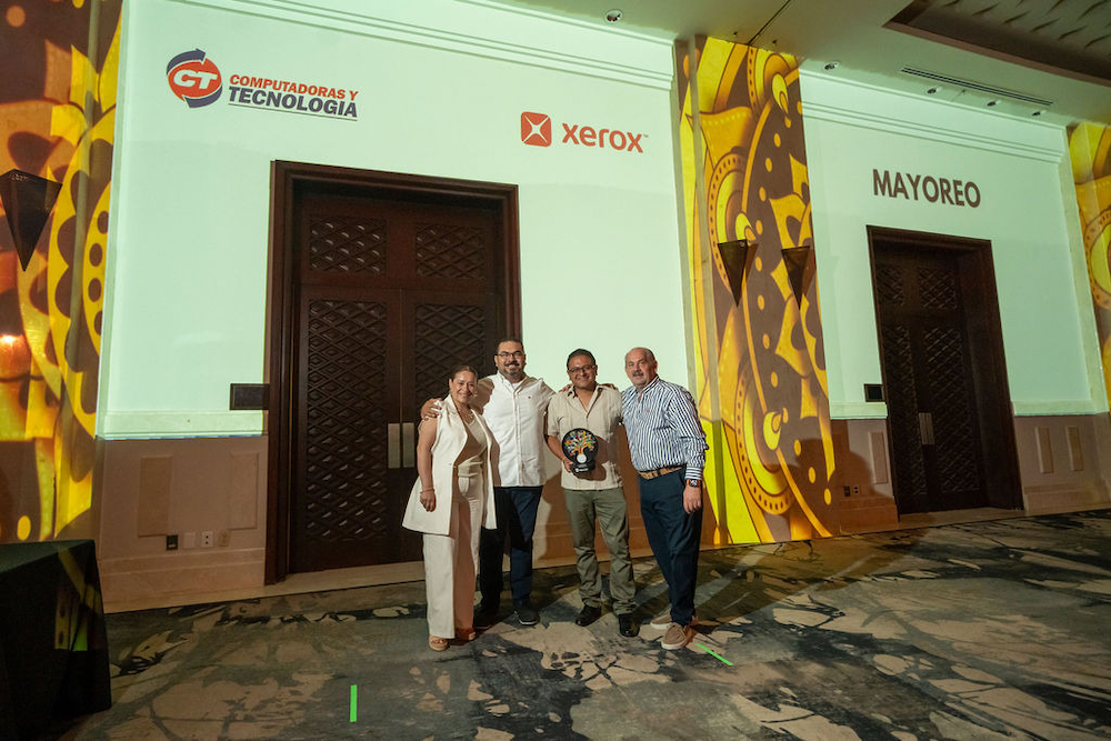 Fotografía en plano general de cuatro directivos de CT Internacional y Xerox posando sonrientes abrazados en el escenario de un salón de eventos con iluminación decorativa, debajo de las proyecciones de los logotipos institucionales de CT Computadoras y Tecnología, Xerox y la palabra Mayoreo.