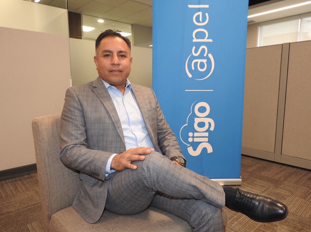 Fotografía en plano medio de Carlos Antonio Ibáñez, abogado especialista en materia fiscal y financiera, vistiendo traje gris a cuadros sobre camisa azul claro, sentado sonriente frente a la mampara corporativa de Siigo Aspel.