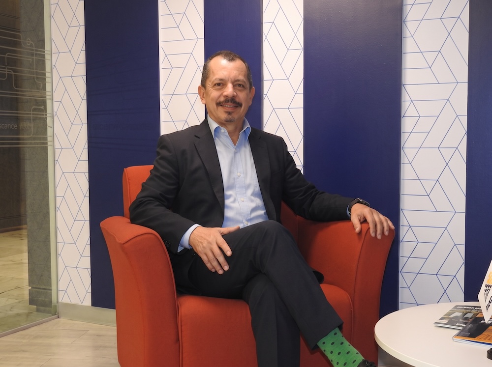 Fotografía en plano medio de Carlos Arochi, Director de Panduit Latam, vistiendo traje oscuro con camisa azul, sentado sonriente con las piernas cruzadas en un sillón rojo corporativo frente a un muro con patrones geométricos azules.