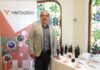 Rentabilidad y nuevas oportunidades para el canal con las baterías de litio de Verbatim Fotografía de plano medio de Carlos Valdés, Country Manager de Verbatim México, posando junto a una mesa de exhibición con productos de la marca y un banner promocional corporativo.