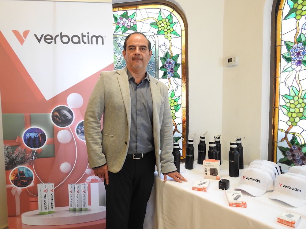 Carlos Valdés impulsa la estrategia de abasto directo y rentabilidad para el canal de distribución en México Fotografía de plano medio de Carlos Valdés, Country Manager de Verbatim México, posando junto a una mesa de exhibición con productos de la marca y un banner promocional corporativo.