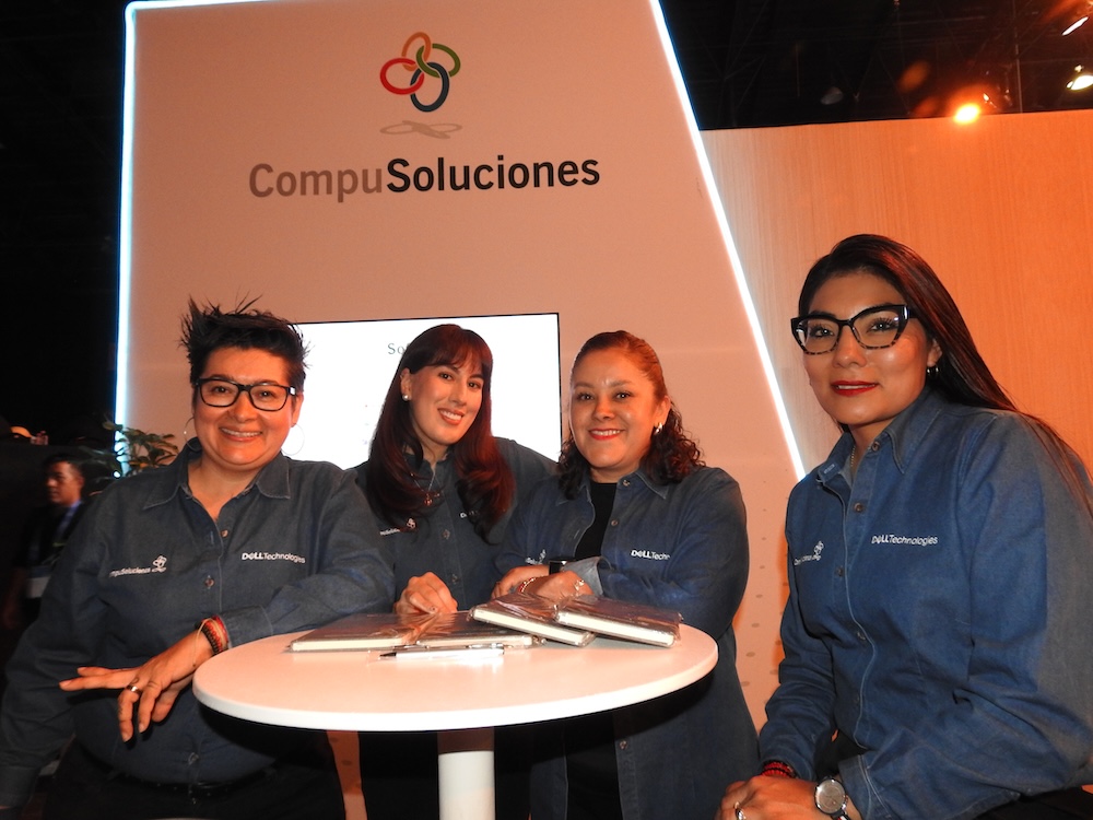 Fotografía de plano medio de cuatro ejecutivas de la célula de Dell Technologies en CompuSoluciones vistiendo camisas de mezclilla con los logotipos corporativos bordados, posando sonrientes alrededor de una mesa alta blanca en su estand de exhibición.