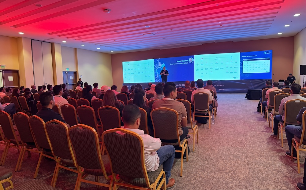 Fotografía desde la parte trasera del salón lleno de asistentes sentados frente al escenario, iluminado con luces de color rojo, donde un ponente expone un diagrama técnico en la pantalla gigante.