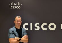 Cisco y la nueva era de la seguridad autónoma como oportunidad dorada para el canal Fotografía de retrato en plano medio de Craig Connors, CTO de infraestructura y seguridad de Cisco, vistiendo playera oscura y reloj inteligente, posando sonriente con los brazos cruzados frente a un muro corporativo con el logotipo de CISCO.