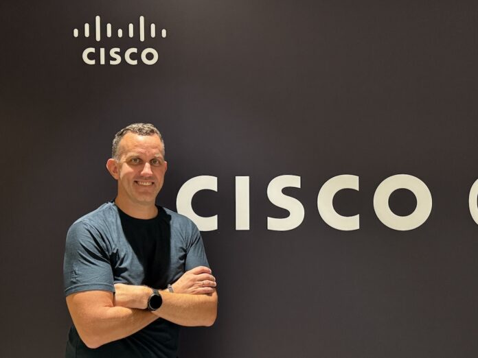 Craig Connors impulsa la adaptación del modelo Zero Trust y la integración de red con ciberseguridad Fotografía de retrato en plano medio de Craig Connors, CTO de infraestructura y seguridad de Cisco, vistiendo playera oscura y reloj inteligente, posando sonriente con los brazos cruzados frente a un muro corporativo con el logotipo de CISCO.
