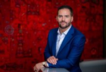 David Jiménez asume como CEO Global de Softtek para impulsar una estrategia AI-First Fotografía de retrato ejecutivo en plano medio de David Jiménez, CEO Global de Softtek, luciendo un saco color azul marino sobre una camisa blanca abierta sin corbata, posando sonriente apoyado en una superficie lisa frente a una pared decorada con arte gráfico en tonos rojos.