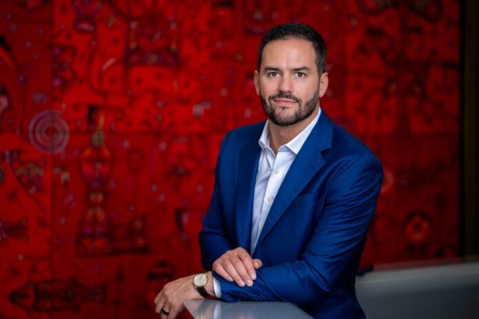Fotografía de retrato ejecutivo en plano medio de David Jiménez, CEO Global de Softtek, luciendo un saco color azul marino sobre una camisa blanca abierta sin corbata, posando sonriente apoyado en una superficie lisa frente a una pared decorada con arte gráfico en tonos rojos.