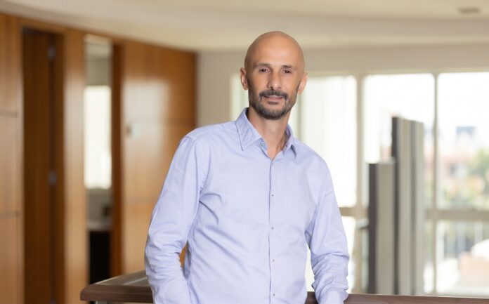 Fotografía de retrato en plano medio de Diego Imperio, Presidente y CEO de Ricoh LATAM, vistiendo una camisa de vestir azul claro y posando sonriente y apoyado en un barandal de madera dentro de un entorno corporativo iluminado.